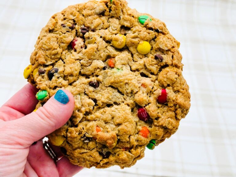 A hand with blue nail polish holds a large, round oatmeal cookie with colorful candy-coated chocolates and chocolate chips in it. The background is a light, plaid surface.