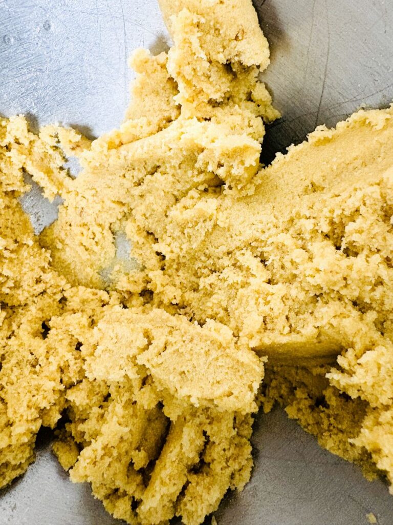 A close-up of yellowish cookie dough being mixed in a metal bowl, showing its crumbly and soft texture.