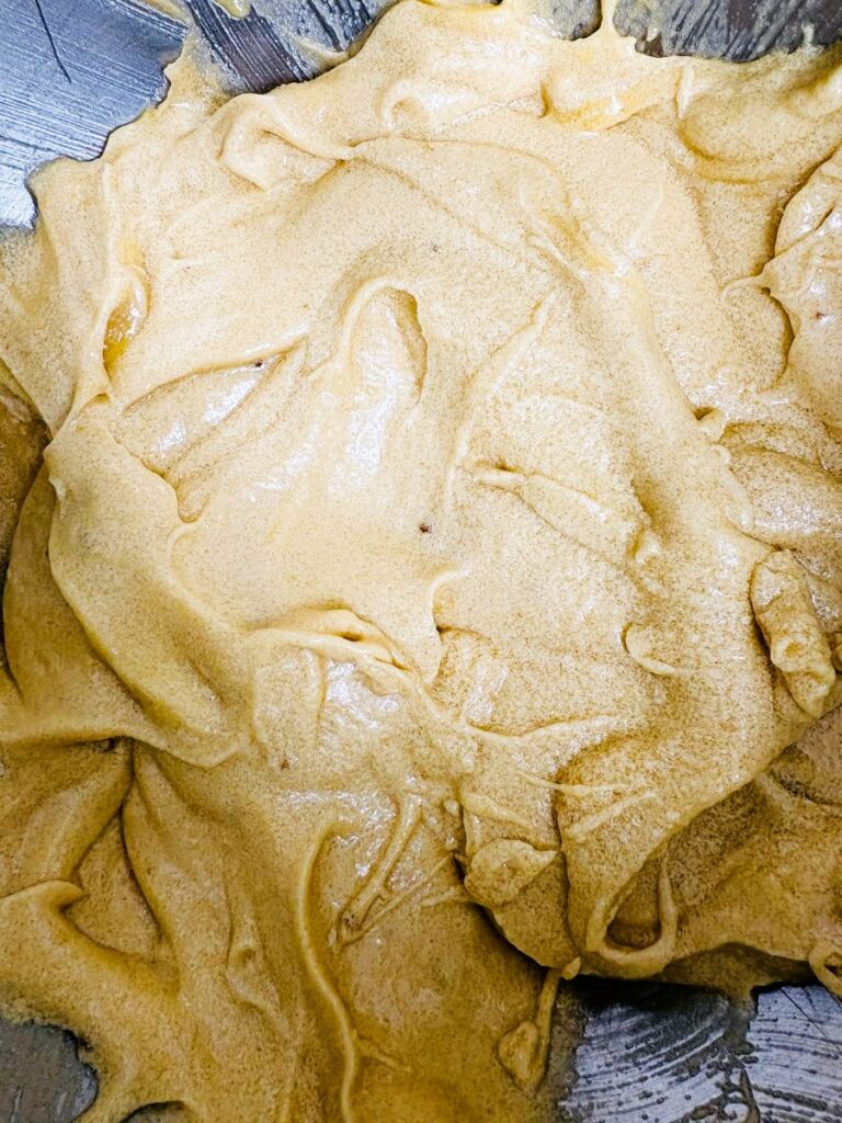 Thick, creamy light brown batter spread across a metal mixing bowl, showing a smooth and slightly fluffy texture with visible swirls and folds.