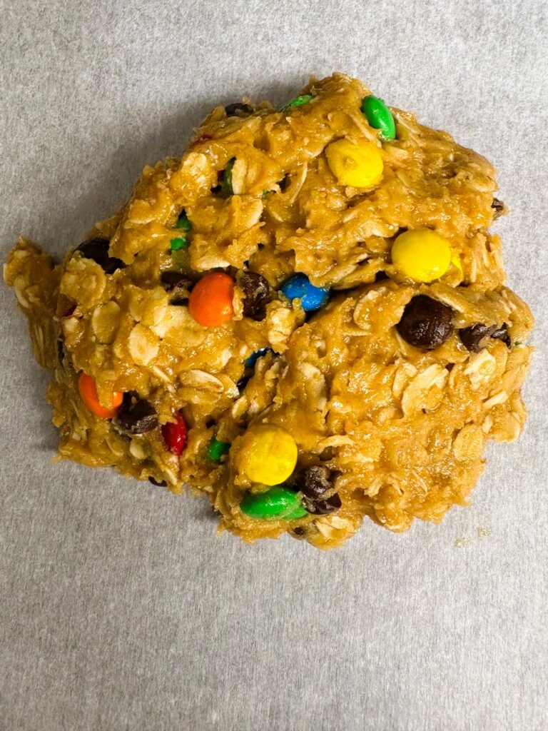 A mound of unbaked cookie dough with oats, colorful candy-coated chocolates, and chocolate chips on a sheet of parchment paper.