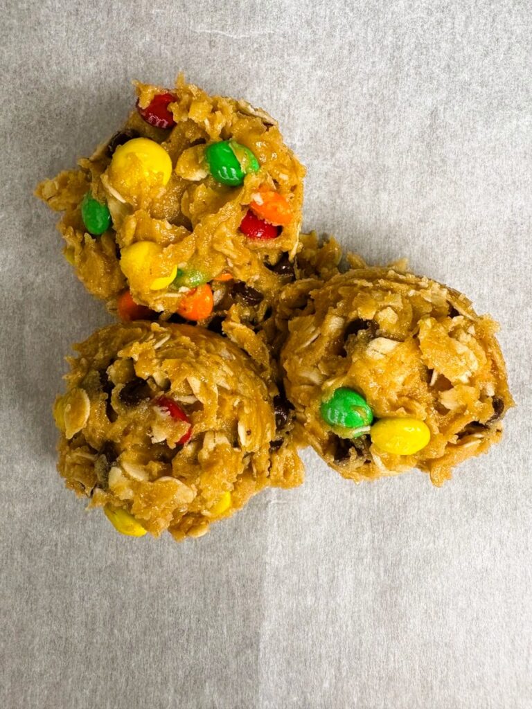 Three unbaked cookie dough mounds with rolled oats, chocolate chips, and colorful candy-coated chocolates on a sheet of parchment paper.