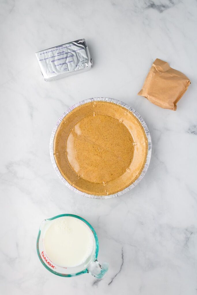 A graham cracker pie crust in a foil pan sits on a marble surface, surrounded by a block of cream cheese, a brown packet of pudding mix, and a glass measuring cup filled with milk.