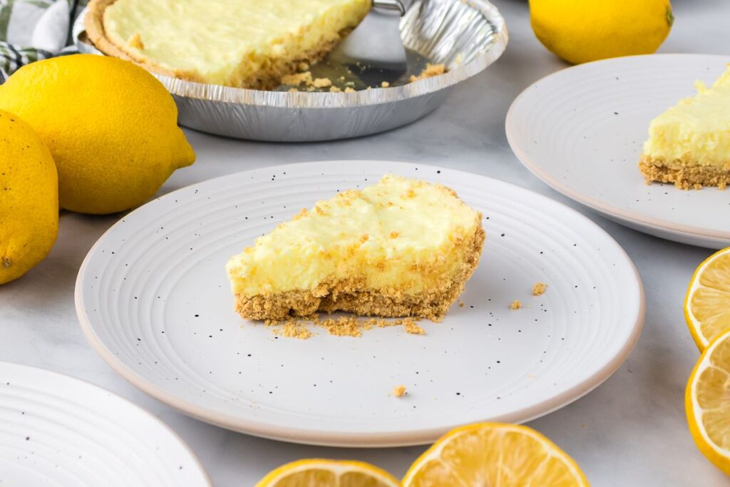 A slice of lemon pie with a crumbly crust sits on a white plate, surrounded by fresh whole and sliced lemons; the remaining pie is in a foil tin in the background.