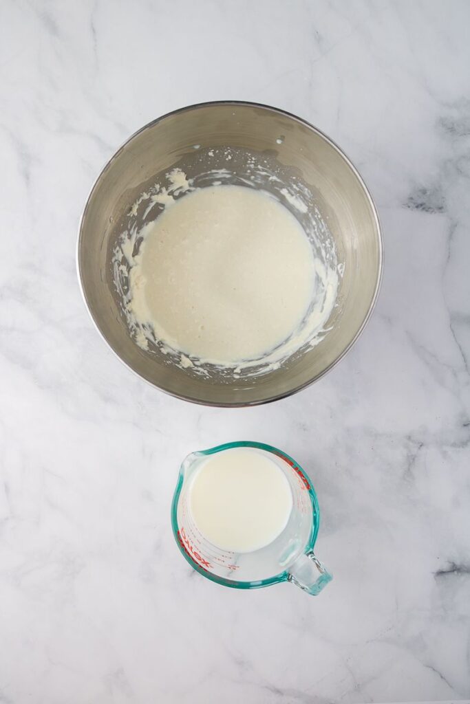 A metal mixing bowl with a small amount of thick white batter inside sits on a marble surface next to a glass measuring cup filled with milk.