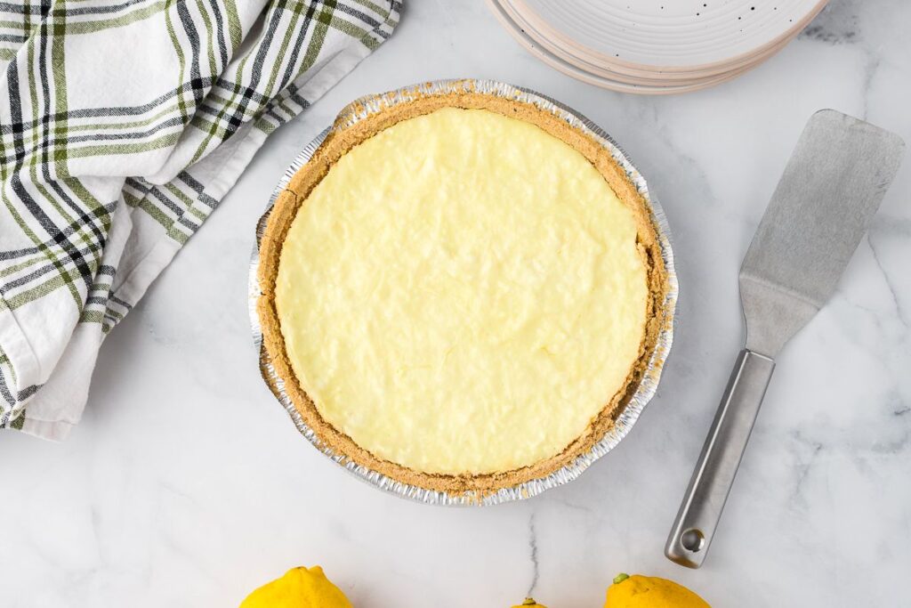 A lemon pie with a graham cracker crust sits on a marble surface next to a metal spatula, a striped kitchen towel, two lemons, and a stack of plates.
