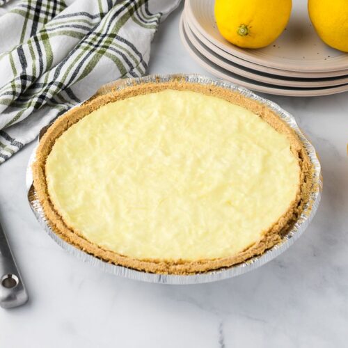 A lemon pie with a graham cracker crust in a foil pan sits on a marble surface. Nearby are three whole lemons, stacked plates, a pie server, and a striped kitchen towel.