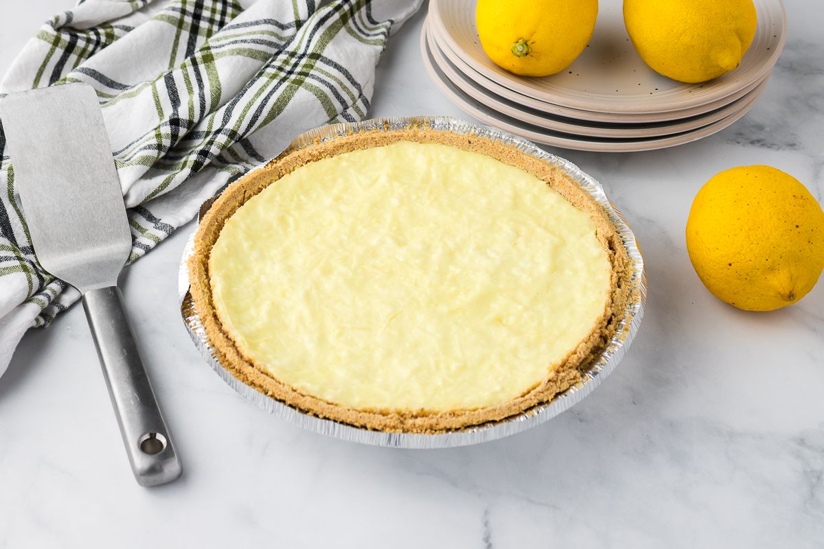 A lemon pie with a graham cracker crust in a foil pan sits on a marble surface. Nearby are three whole lemons, stacked plates, a pie server, and a striped kitchen towel.