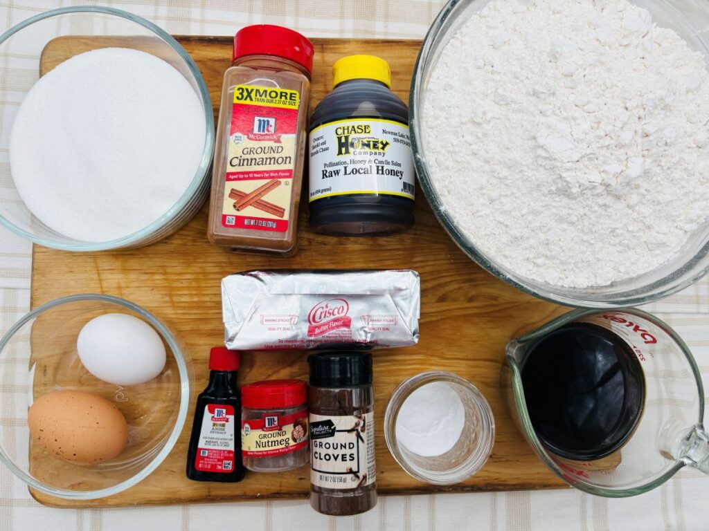 A wooden board holds baking ingredients: a bowl of sugar, eggs, nutmeg, cloves, cinnamon, honey, flour, baking soda, shortening, vanilla extract, and a measuring cup of molasses on a checkered tablecloth.