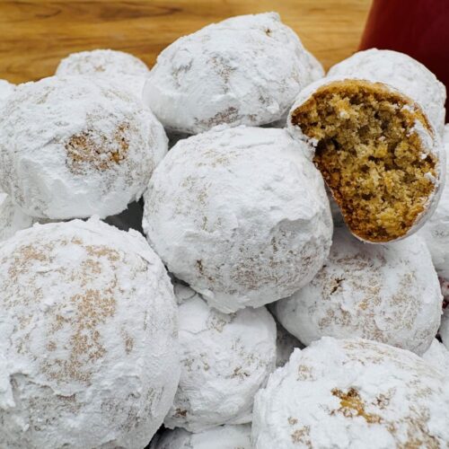 A pile of round cookies coated in powdered sugar is shown on a plate. One cookie is broken in half, revealing a crumbly, brown interior. The background includes a wooden surface and a red item.