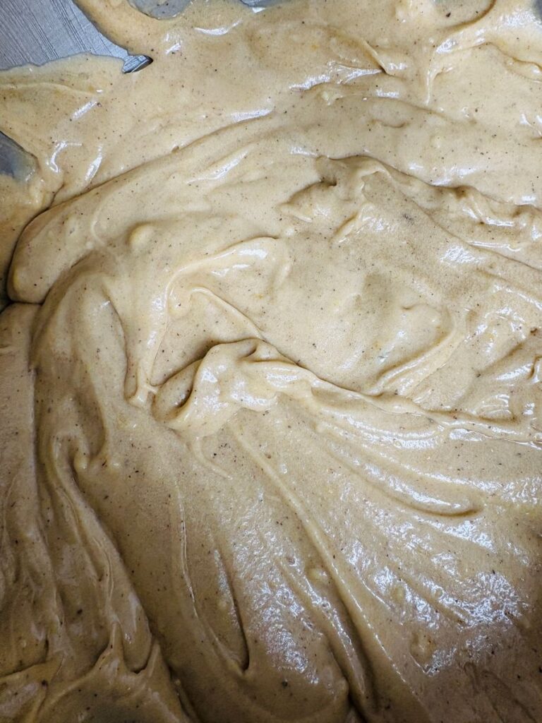 A close-up of thick, light brown batter or dough spread unevenly across a flat surface, with visible swirls and peaks in the texture.
