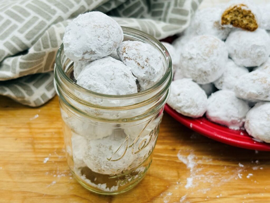 A glass jar filled with round cookies coated in powdered sugar sits on a wooden surface, with more sugar-dusted cookies on a red plate in the background. A cloth napkin is partially visible beside the jar.