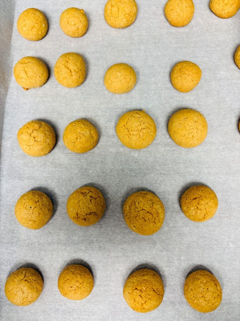 Rows of round, golden-brown cookies are arranged on a sheet of parchment paper, viewed from above. The cookies are evenly spaced and appear freshly baked.