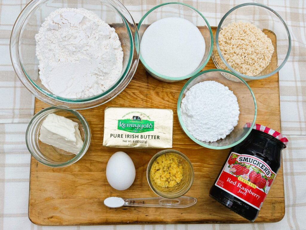 Top view of baking ingredients on a wooden board, including flour, sugar, brown sugar, powdered sugar, butter, cream cheese, an egg, lemon zest, salt, and a jar of red raspberry jam.