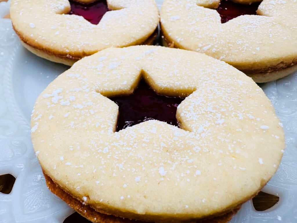 Three round cookies with powdered sugar on top and a star-shaped cutout in the center, revealing red jam filling, are displayed on a white decorative plate.