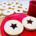Two Linzer cookies with star-shaped jam centers on a red plate, next to a red mug. More cookies are visible in the background on a tray, all dusted with powdered sugar.