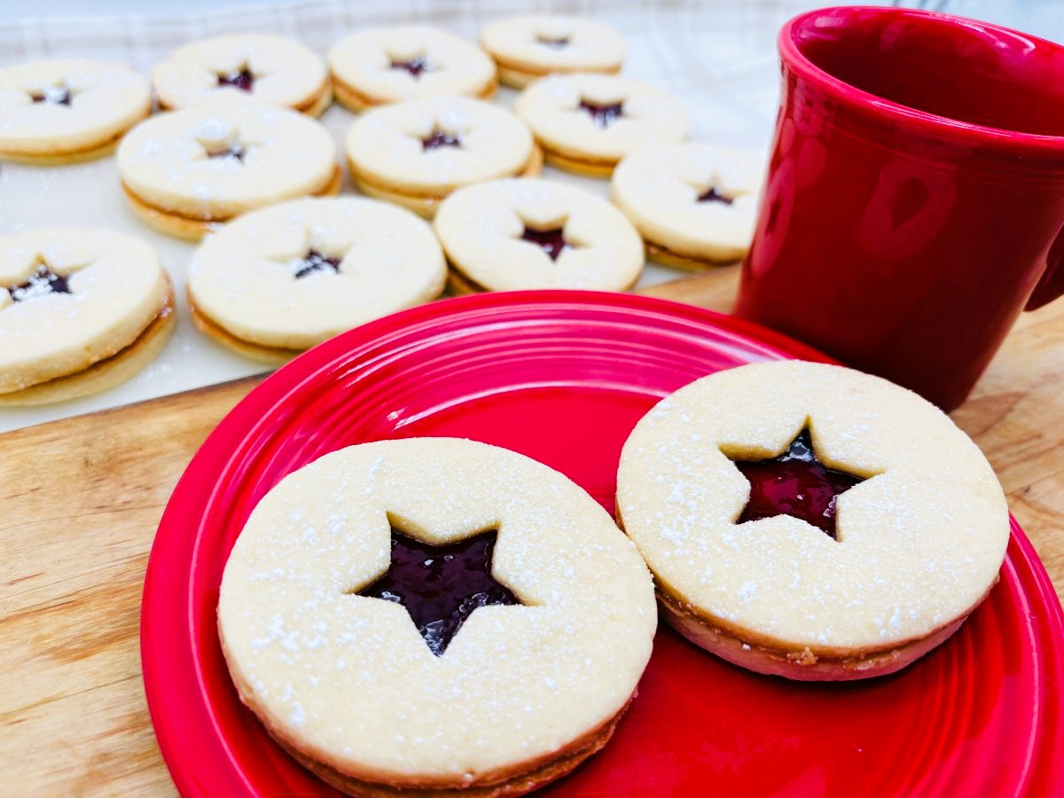 Two Linzer cookies with star-shaped jam centers on a red plate, next to a red mug. More cookies are visible in the background on a tray, all dusted with powdered sugar.