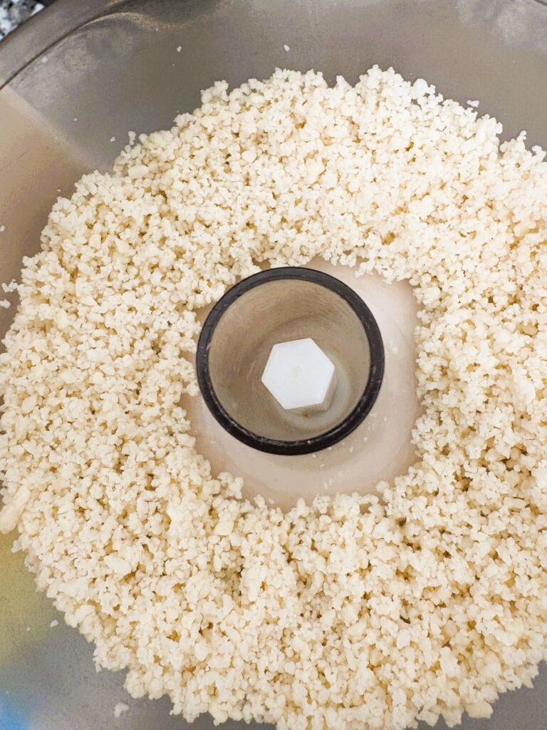 Finely ground almonds resembling rice grains inside a food processor bowl, with the blade attachment visible in the center.