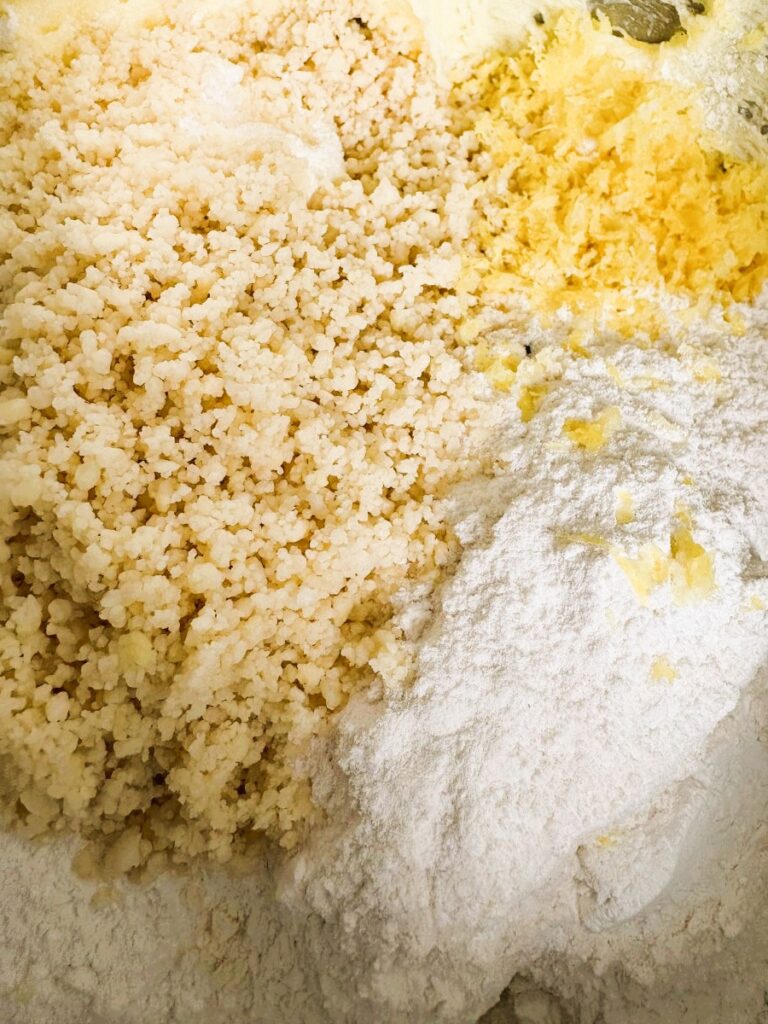 A close-up of a baking mixture, showing granulated sugar, flour, and softened butter partially blended together in a bowl. The ingredients are in the process of being mixed for baking.