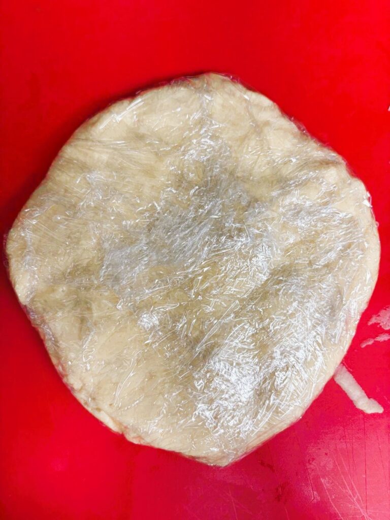 A round disc of dough wrapped in plastic wrap rests on a bright red surface, ready for chilling or storage.