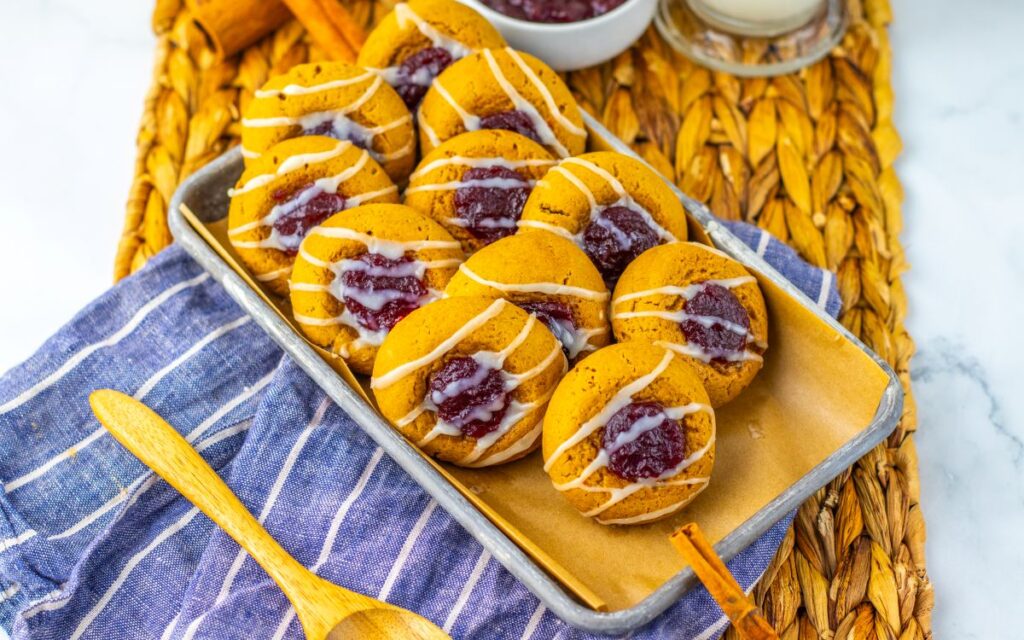 A tray of soft cookies topped with white icing drizzle and a dollop of red jam, set on parchment paper over a blue striped cloth with a wooden spoon, atop a woven placemat.