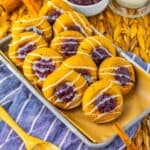 A tray of soft cookies topped with white icing drizzle and a dollop of red jam, set on parchment paper over a blue striped cloth with a wooden spoon, atop a woven placemat.