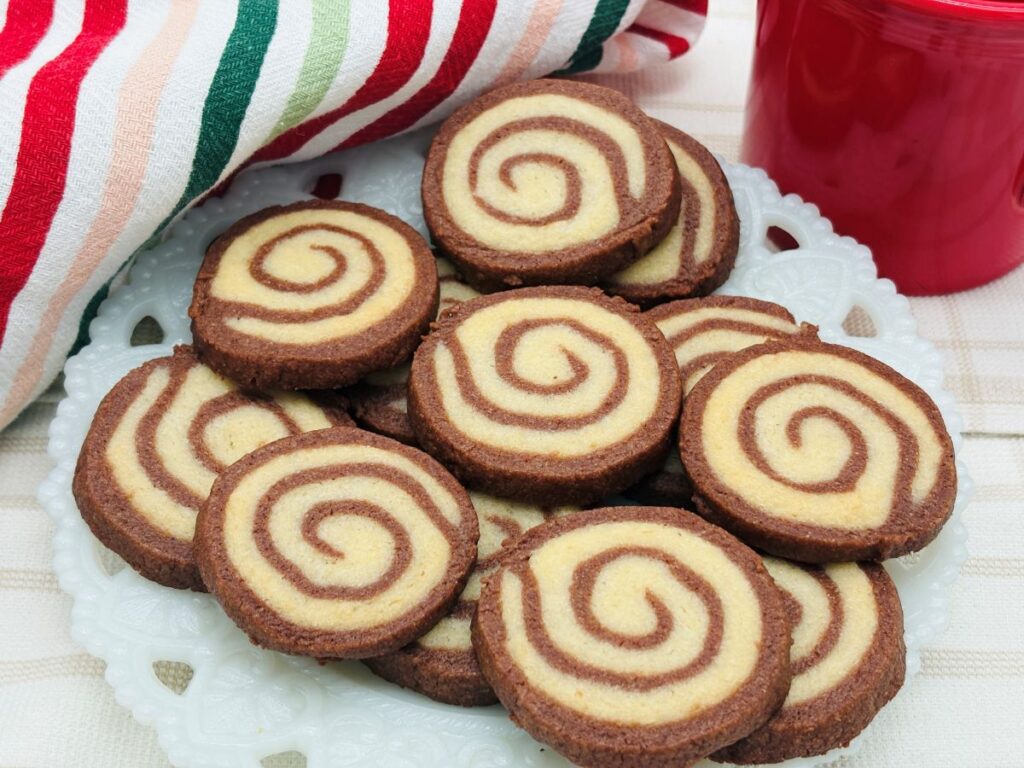 A plate of spiral chocolate and vanilla cookies sits on a white decorative dish, with a red cup and a striped cloth in the background.