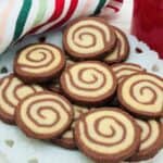 A plate of spiral chocolate and vanilla cookies sits on a white decorative dish, with a red cup and a striped cloth in the background.