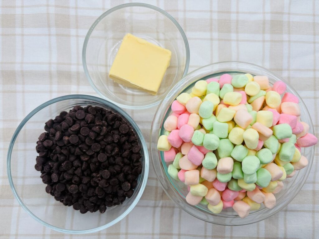 Three glass bowls on a plaid surface: one with chocolate chips, one with a square of butter, and a large one filled with colorful mini marshmallows.