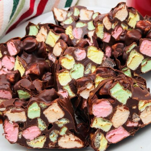 A plate filled with pieces of chocolate-covered dessert bars packed with colorful mini marshmallows, with a red and white striped cloth in the background.