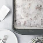 A frosted sheet cake in a square baking pan sits on a marble surface next to a cake server, a plate with two forks, and a green-striped kitchen towel.