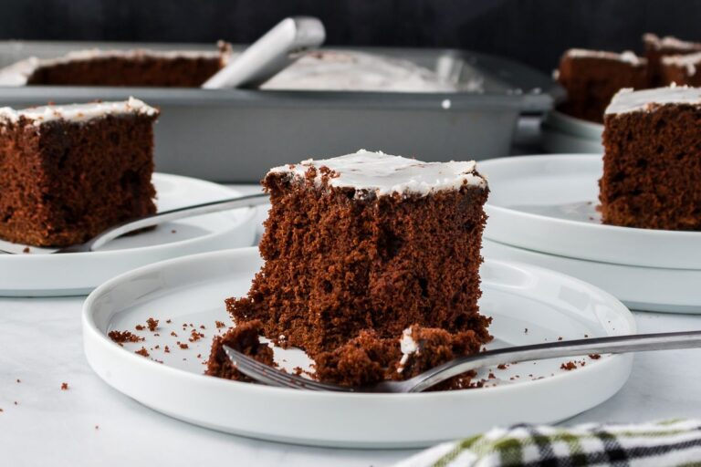 A slice of chocolate cake with white icing sits on a white plate, partially eaten with a fork. More slices and a baking pan with cake are visible in the background. Crumbs are scattered on the plates.