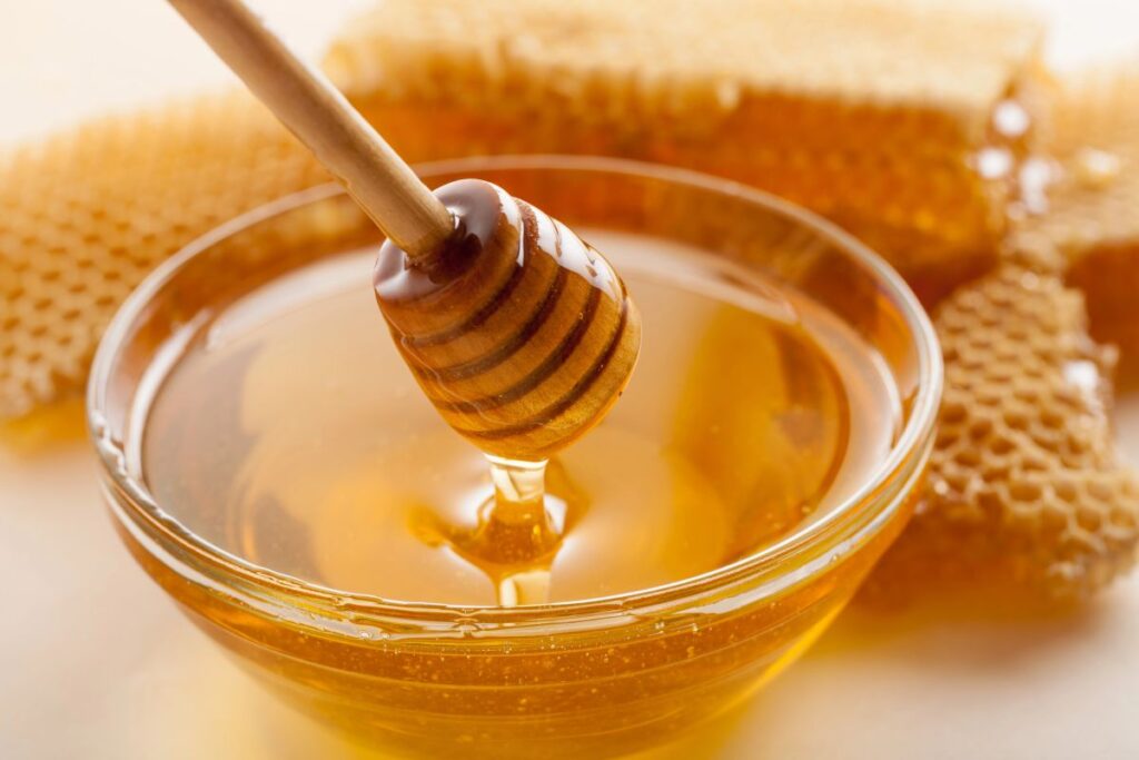 A wooden honey dipper drizzling golden honey into a glass bowl, with pieces of honeycomb in the background.