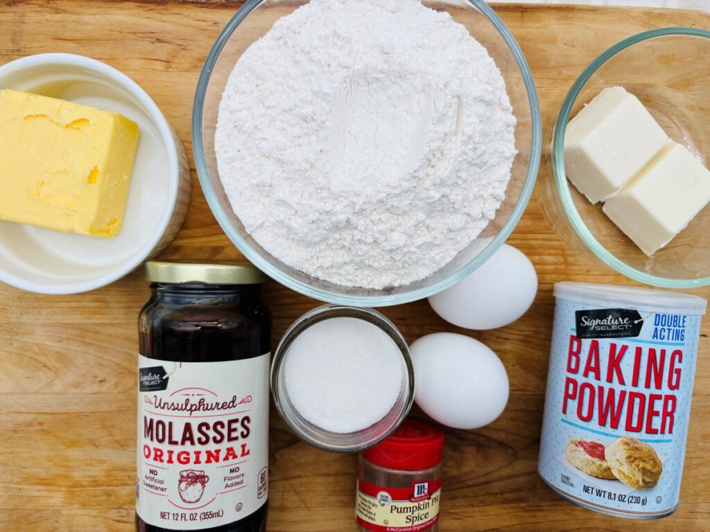 Baking ingredients on a wooden surface, including butter, flour, eggs, baking powder, sugar, molasses, pumpkin pie spice, and cream cheese. Each item is in a bowl, jar, or container.