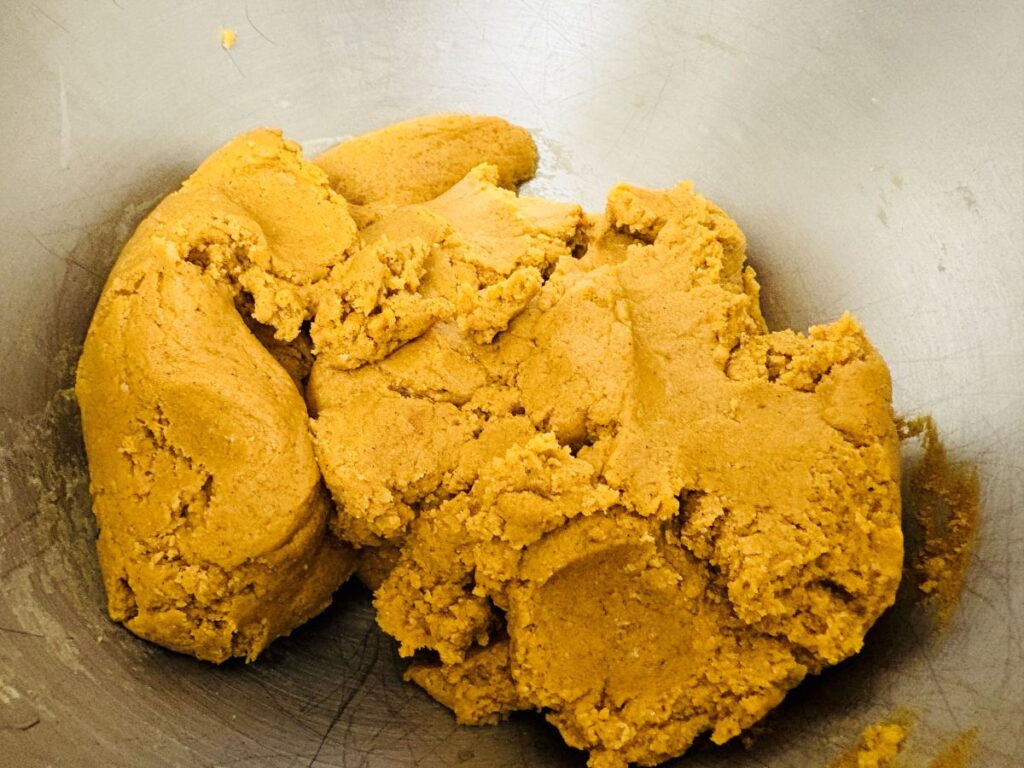 A close-up of raw cookie dough with a light brown color, resting in a large metallic mixing bowl. The dough appears soft and slightly crumbly in texture.
