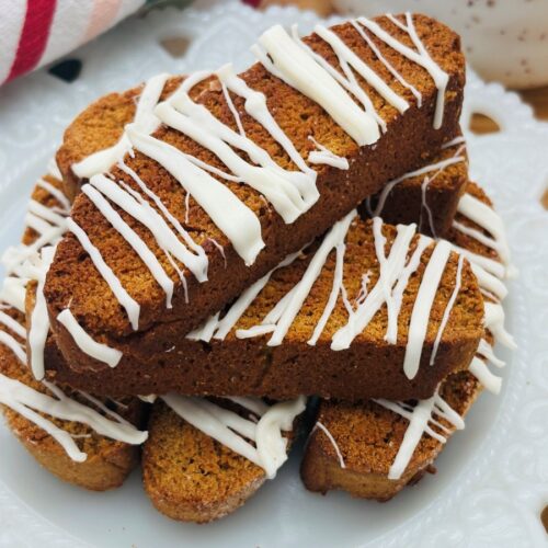 A plate of biscotti cookies drizzled with white icing is arranged on a decorative white plate. The cookies are golden brown and stacked neatly, with a striped towel visible in the background.
