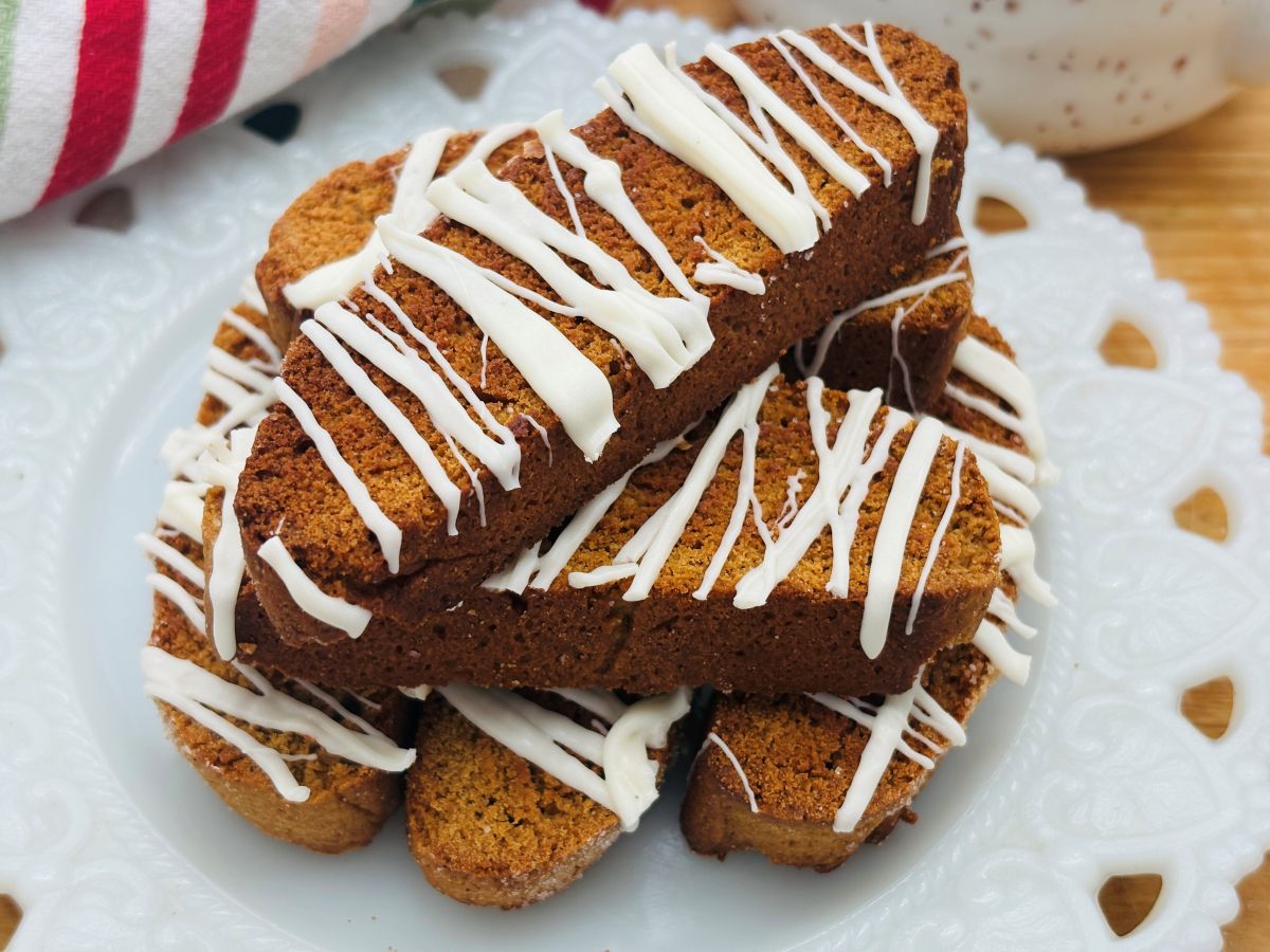 A plate of biscotti cookies drizzled with white icing is arranged on a decorative white plate. The cookies are golden brown and stacked neatly, with a striped towel visible in the background.