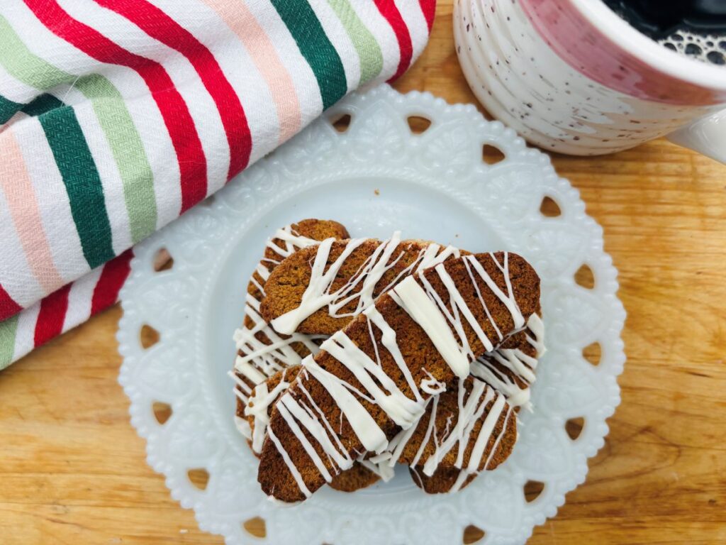A plate of gingerbread cookies drizzled with white icing sits on a wooden surface next to a striped cloth and a mug of coffee.
