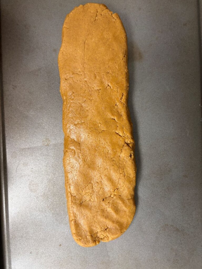 A large, unevenly shaped, brown slab of dough is spread on a metal baking sheet, ready to be baked. The dough has a rough, cracked surface and appears homemade.
