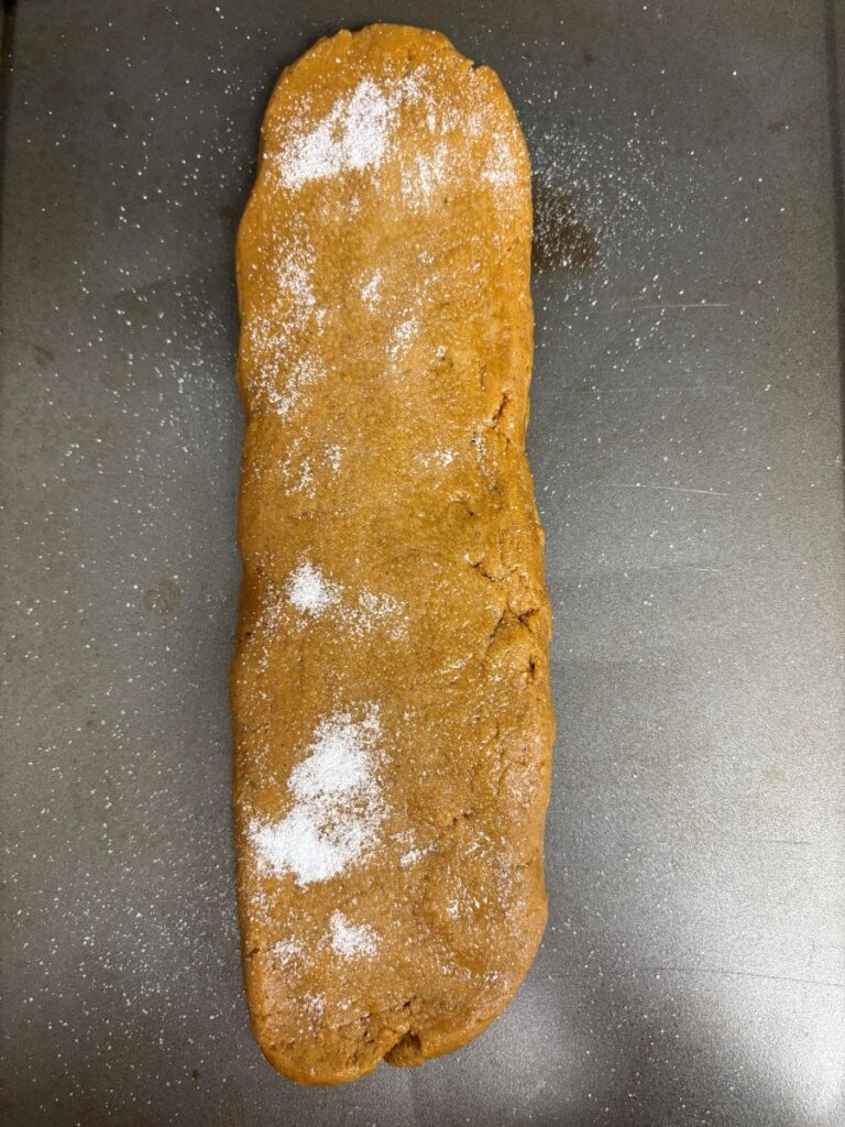 A long, rectangular loaf of brown dough with a lightly cracked surface is sprinkled with white sugar and placed on a metal baking sheet.