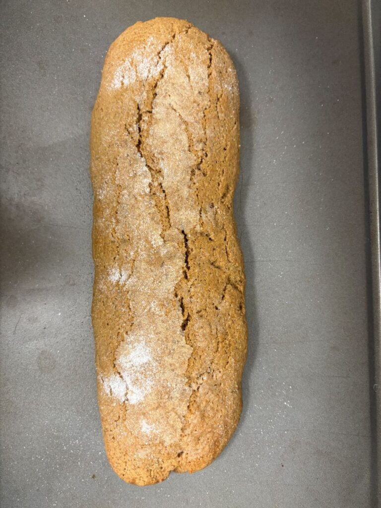A single loaf of rustic, brown gingerbread with a cracked top and a dusting of flour sits on a gray baking sheet.