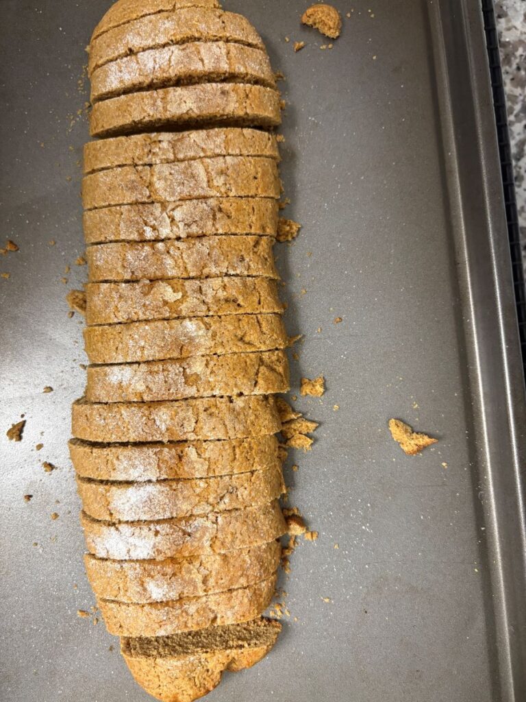 A loaf of biscotti sliced into even pieces on a baking tray, with a few crumbs scattered around. The biscotti appears golden brown and lightly dusted with sugar.