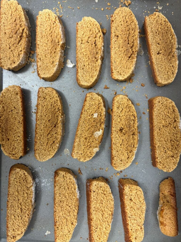 A baking tray with 15 evenly spaced slices of golden-brown biscotti, arranged in rows. The biscotti are slightly crumbly with some visible flour on the tray.