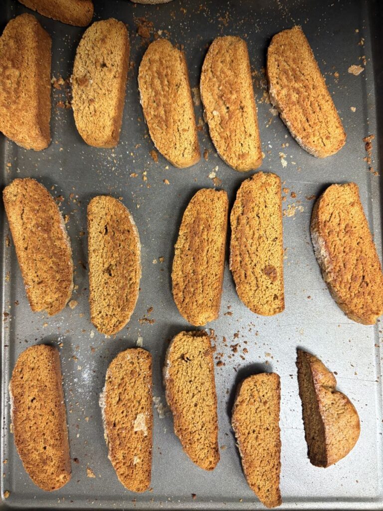 A baking sheet covered with rows of golden-brown biscotti, some with visible crumbs and rough, crunchy edges. The biscotti are arranged haphazardly, suggesting they have just been baked.