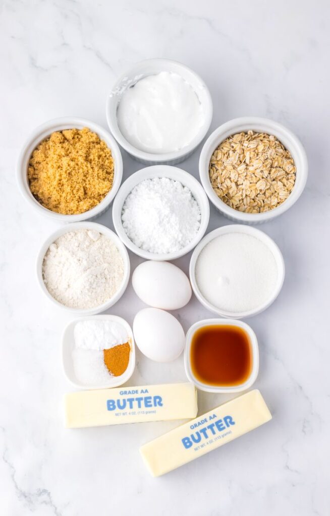 An assortment of baking ingredients in small bowls and on a marble surface, including brown sugar, powdered sugar, oats, flour, eggs, white sugar, baking soda, salt, cinnamon, vanilla extract, and two sticks of butter.