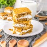 Three oatmeal cream pies are stacked on a white plate, filled with white cream, with a glass of milk, cinnamon sticks, and wooden spoons nearby on a wooden tray.
