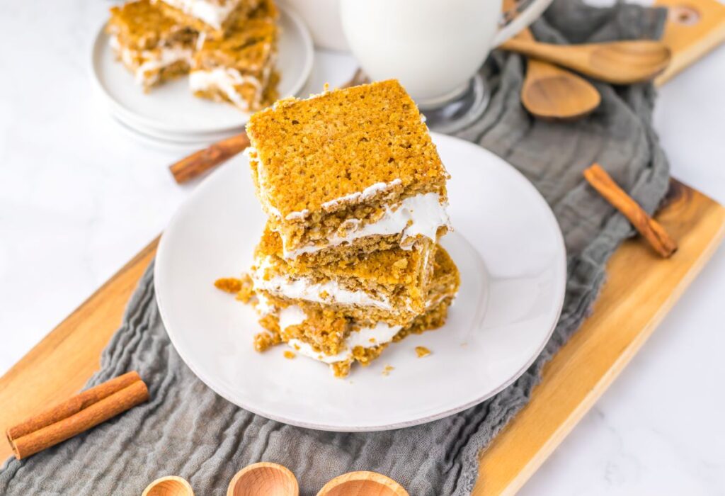 Three oatmeal cream pie bars with white cream filling are stacked on a white plate. A glass of milk, cinnamon sticks, wooden spoons, and more bars are in the background on a wooden tray with a gray cloth.