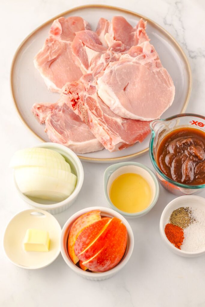 A plate with raw pork chops, a bowl of sliced onions, a small bowl of butter, apple slices, a cup of barbecue sauce, a small bowl of apple cider vinegar, and a bowl of spices on a white surface.