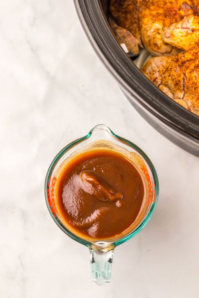 A glass measuring cup filled with thick, reddish-brown barbecue sauce sits on a white surface next to a slow cooker containing seasoned, cooked chicken.