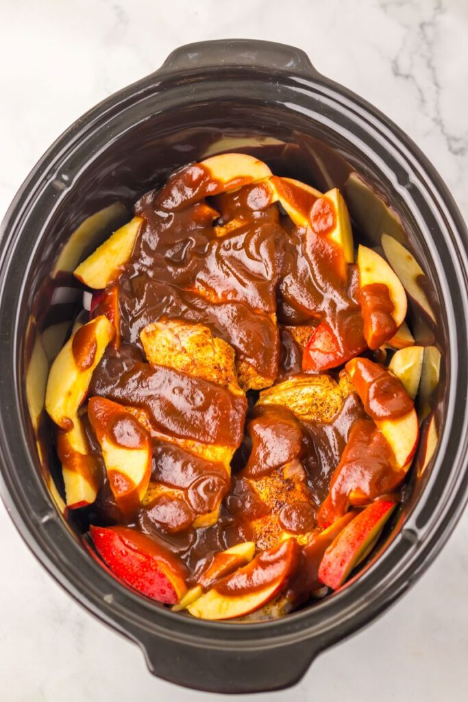 Sliced apples and pieces of chicken are topped with barbecue sauce inside a black slow cooker, ready to be cooked. The cooker sits on a white marble surface.