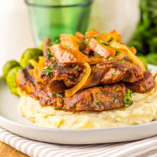 A plate of mashed potatoes topped with braised beef strips, sautéed onions, and garnished with parsley, accompanied by Brussels sprouts, served on a striped napkin.